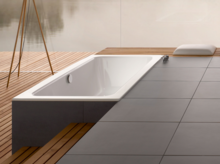 Baignoire rectangulaire en acier émaillé, partiellement encastrée dans un sol en bois, avec entourage moderne en carrelage.