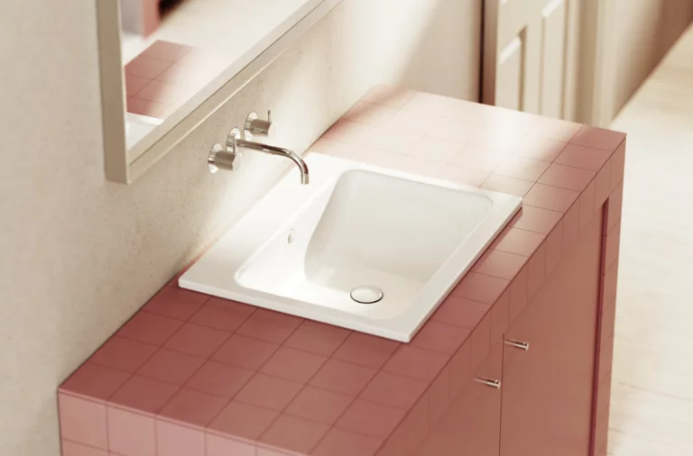 Умывальник с белой чашей на розовой плитке, настенный смеситель и зеркало на заднем плане.