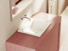 Умывальник с белой чашей на розовой плитке, настенный смеситель и зеркало на заднем плане.