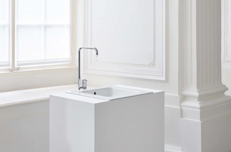 Прямоугольная стальная эмалированная раковина на белой столешнице с краном, перед светлым окном и колонной.