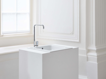 Прямоугольная стальная эмалированная раковина на белой столешнице с краном, перед светлым окном и колонной.