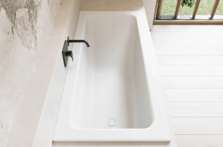 Weiße Badewanne in heller Nische mit schwarzer Wandarmatur und hellem Boden am Fenster.