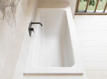 Weiße Badewanne in heller Nische mit schwarzer Wandarmatur und hellem Boden am Fenster.