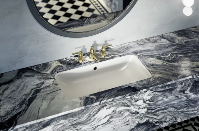 Rectangular white inset washbasin in marbled countertop with gold faucet and round mirror above.