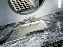 Rectangular white inset washbasin in marbled countertop with gold faucet and round mirror above.