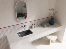 Helles Badezimmer mit dunkelgrauem Waschtisch, silberfarbenen Wandarmaturen, ovalem Spiegel, blauer Vase und gefliester Ablage.