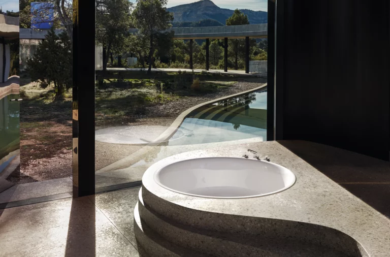 Vasca da bagno rotonda in acciaio smaltato inserita in piano in pietra organica, grande finestra con vista sul verde.
