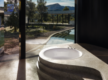 Vasca da bagno rotonda in acciaio smaltato inserita in piano in pietra organica, grande finestra con vista sul verde.