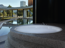 Vasca da bagno rotonda in acciaio smaltato, incassata a gradini, con rubinetteria, vista sull’esterno attraverso ampia vetrata.