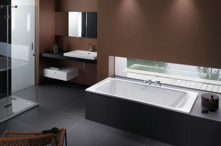 Rechthoekig bad van staal-email, wastafel op inbouwblad, moderne douche, donkere tegels, groot raam.