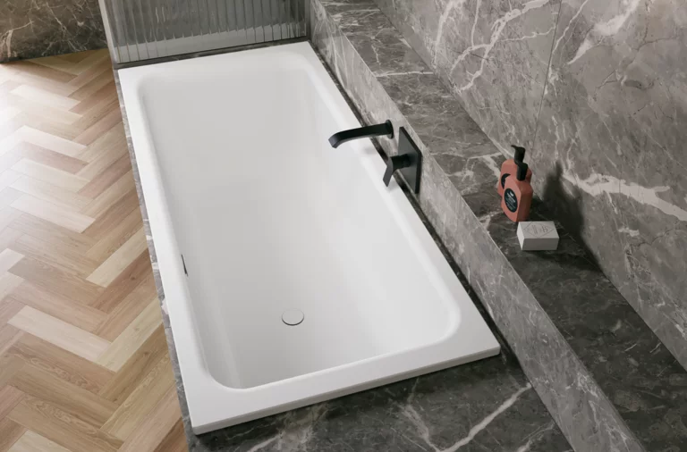 Weiße Badewanne in grauer Nische mit schwarzer Armatur, Ablage und hellem Fischgrätboden.