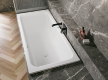 Weiße Badewanne in grauer Nische mit schwarzer Armatur, Ablage und hellem Fischgrätboden.