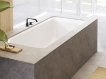 Vasca da bagno incassata rettangolare con rivestimento grigio, rubinetto nero e luce naturale da destra.