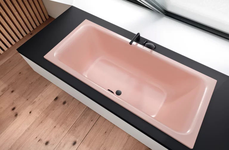 Rechteckige, eingebaute Badewanne in zartem Rosé, mit schwarzer Armatur, vor Fenster und auf hellem Holzboden.