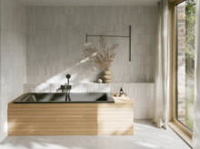 Dunkle Badewanne mit gerillter Front in hellem Bad mit weißen Fliesen, Fenster und Vase.