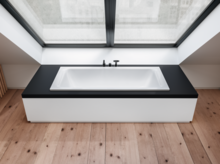 Vasca da bagno rettangolare in acciaio smaltato, integrata in piano nero, sotto grande finestra, ambiente moderno con pavimento in legno.