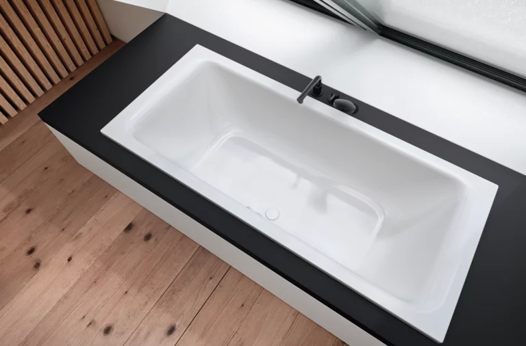 Rechteckige Badewanne aus Stahl-Email, eingebaut in schwarze Fläche, mit Armatur, modernes Design, große Fensterfront.