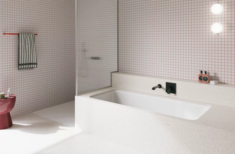 Weiße Badewanne in weiß gefliestem Bad mit Duschbereich, Wandarmatur und rosa Hocker.
