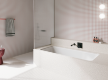 Weiße Badewanne in weiß gefliestem Bad mit Duschbereich, Wandarmatur und rosa Hocker.