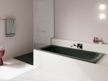 Dunkle Badewanne in hell gefliestem Bad mit Duschbereich, Wandarmatur und rosa Hocker.