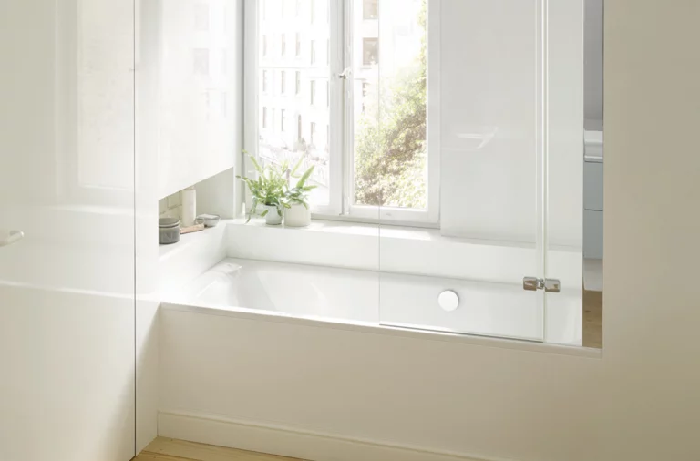 Rectangular steel-enamel bathtub under window, with glass screen, light wall, wooden floor, plants on shelf.