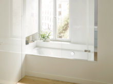 Rectangular steel-enamel bathtub under window, with glass screen, light wall, wooden floor, plants on shelf.