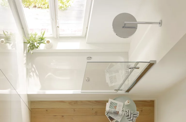 Rectangular steel enamel bathtub under window, glass shower screen beside, round mirror and small side table with magazine.