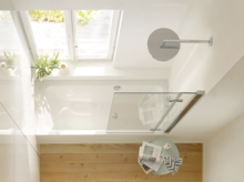 Rectangular steel enamel bathtub under window, glass shower screen beside, round mirror and small side table with magazine.