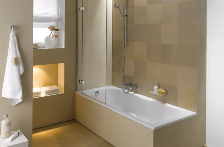 Baignoire rectangulaire en acier émaillé avec paroi de douche, salle de bains carrelée beige, douchette, serviette, niche de rangement.