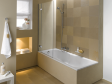 Baignoire rectangulaire en acier émaillé avec paroi de douche, salle de bains carrelée beige, douchette, serviette, niche de rangement.