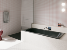 Dunkle Badewanne in hell gefliestem Bad mit Duschbereich, Wandarmatur und rosa Hocker.