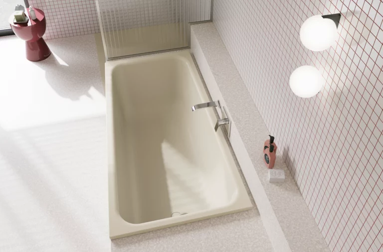 Crèmekleurig bad in lichte badkamer met witte tegels, zilverkleurige kraan, ronde lampen en rood bijzettafeltje.