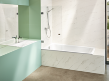 Modernes Bad mit rechteckiger Stahl-Email-Badewanne, Waschbecken auf Einbauplatte, ebenerdigem Boden und heller Wandverkleidung.