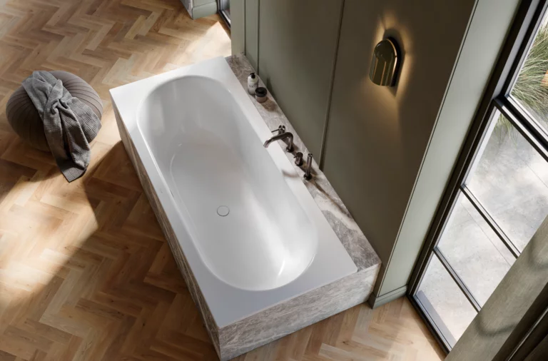 Elegante Badewanne mit Marmorverkleidung vor grünlicher Wand, auf hellem Fischgrätparkett neben bodentiefem Fenster.