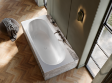 Elegante Badewanne mit Marmorverkleidung vor grünlicher Wand, auf hellem Fischgrätparkett neben bodentiefem Fenster.