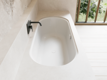 Weiße Badewanne in hellem Bad mit schwarzer Armatur, hellem Boden und Fenster mit Blick ins Grüne.