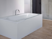 Rectangular steel enamel bathtub with rounded inner edges, installed in modern bathroom with wooden floor.