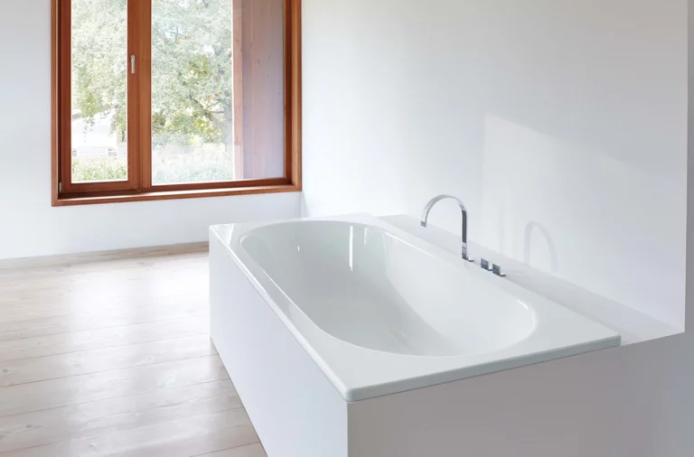 Rectangular steel enamel bathtub in bright setting, built-in with modern fittings and large window in background.