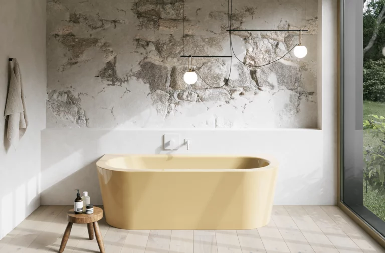 Gelbe freistehende Badewanne in hellem Bad mit Natursteinoptik-Wand, Hocker, Handtuch und großem Fenster.