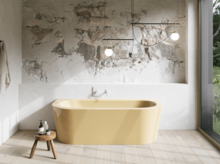 Gelbe freistehende Badewanne in hellem Bad mit Natursteinoptik-Wand, Hocker, Handtuch und großem Fenster.