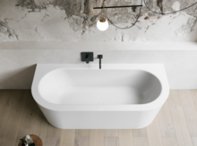 Weiße freistehende Badewanne in hellem Bad mit Natursteinoptik-Wand, schwarzer Armatur und Holzoptik-Boden.