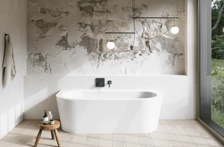 Weiße freistehende Badewanne in hellem Bad mit Natursteinoptik-Wand, Hocker, Handtuch und großem Fenster.