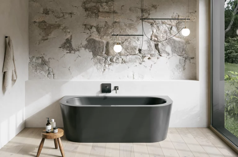 Graue freistehende Badewanne in hellem Bad mit Natursteinoptik-Wand, Hocker, Handtuch und großem Fenster.