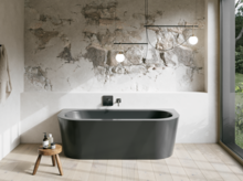 Graue freistehende Badewanne in hellem Bad mit Natursteinoptik-Wand, Hocker, Handtuch und großem Fenster.