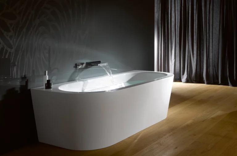 Freistehende ovale Badewanne aus Stahl-Email, Wasser läuft ein, auf Holzfußboden vor dunkler Wand und Vorhang.