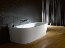 Freistehende ovale Badewanne aus Stahl-Email, Wasser läuft ein, auf Holzfußboden vor dunkler Wand und Vorhang.