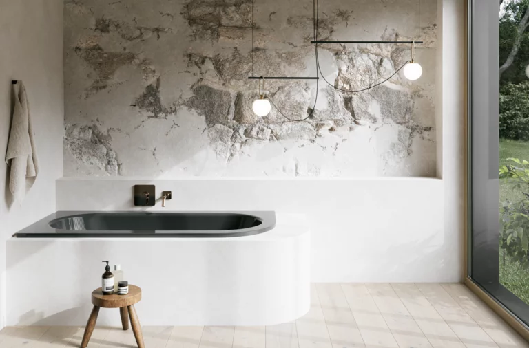 Dunkle Einbau-Badewanne in hellem Bad mit weißer Verkleidung, Natursteinoptik-Wand, Hocker und großem Fenster.