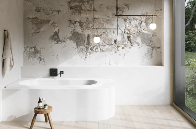 Weiße Einbau-Badewanne in hellem Bad mit Natursteinoptik-Wand, kleinem Hocker und großem Fenster.