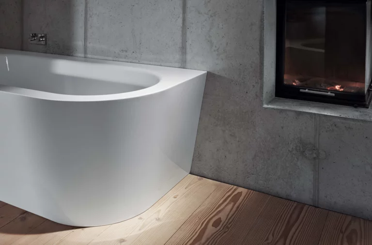 Freistehende Badewanne aus Stahl-Email mit abgerundeter Ecke, vor Betonwand und Kamin, auf Holzfußboden.