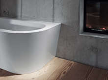 Freistehende Badewanne aus Stahl-Email mit abgerundeter Ecke, vor Betonwand und Kamin, auf Holzfußboden.